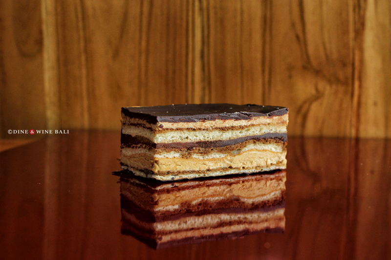 A decadent slice of opera cake with layers of chocolate, coffee, and sponge, displayed on a glossy wooden surface at Poule de Luxe Bali