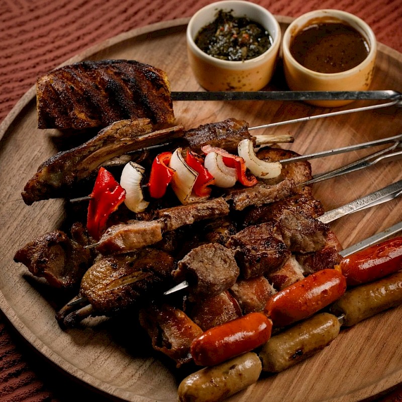 A Brazilian BBQ platter featuring skewered meats, grilled sausages, roasted vegetables, and dipping sauces on a wooden plate at Valle Canggu Padang Linjong