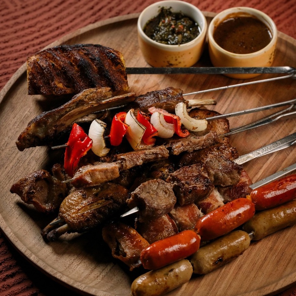 A Brazilian BBQ platter featuring skewered meats, grilled sausages, roasted vegetables, and dipping sauces on a wooden plate at Valle Canggu Padang Linjong